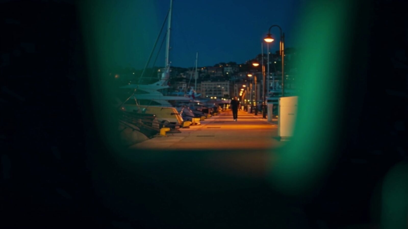 Vue dans des jumelles : le ponton du port à la nuit tombante, une silhouette s'éloigne entre les bateaux