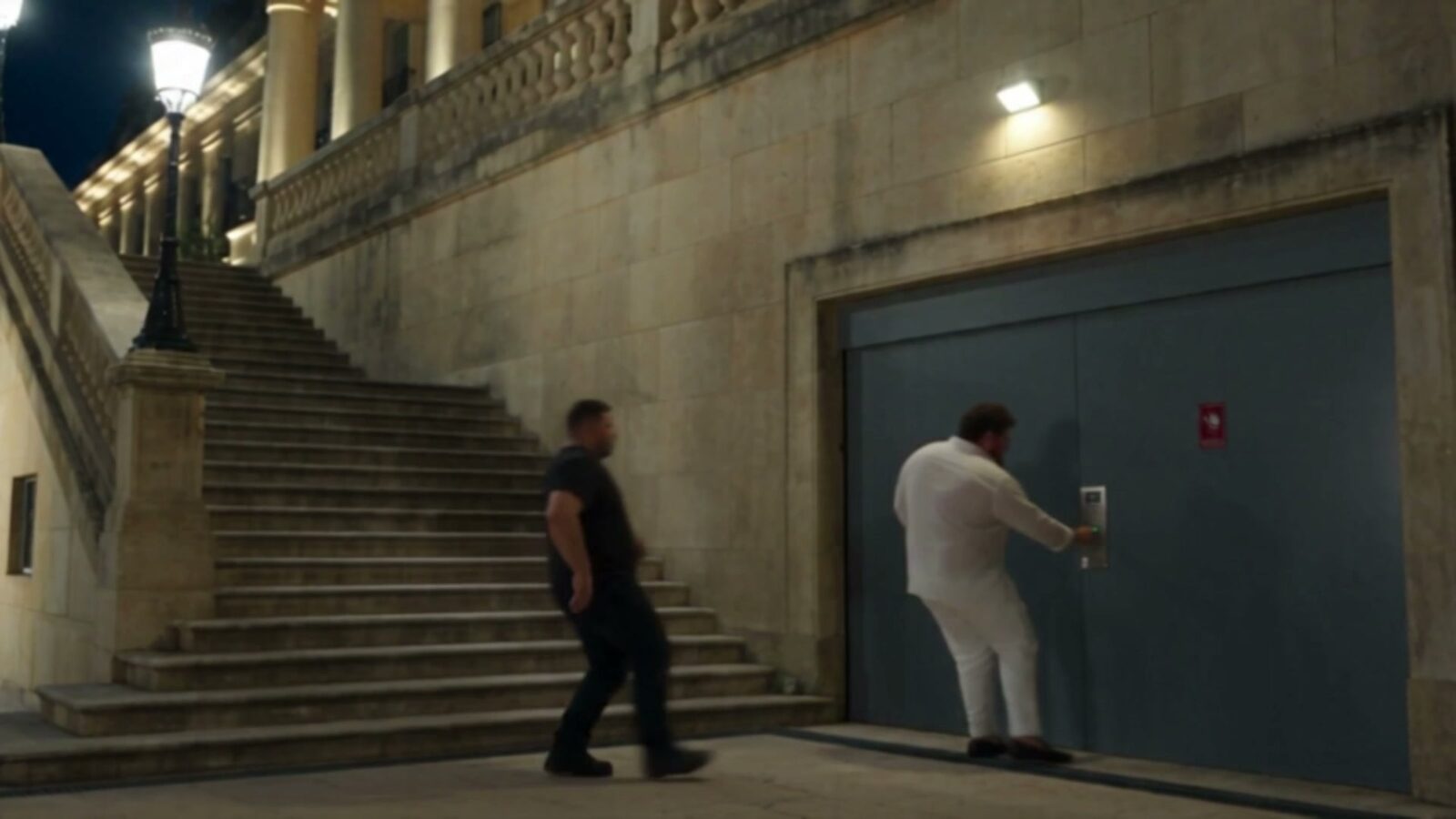 Au pied d'un grand escalier, le milliardaire ouvre une porte de garage pendant qu'un homme le rejoint