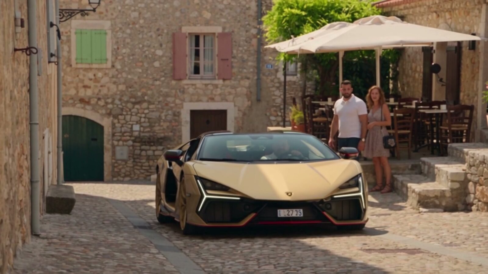 Dernier plan, la Lamborghini dorée garée dans une ruelle de village devant une terrasse au soleil