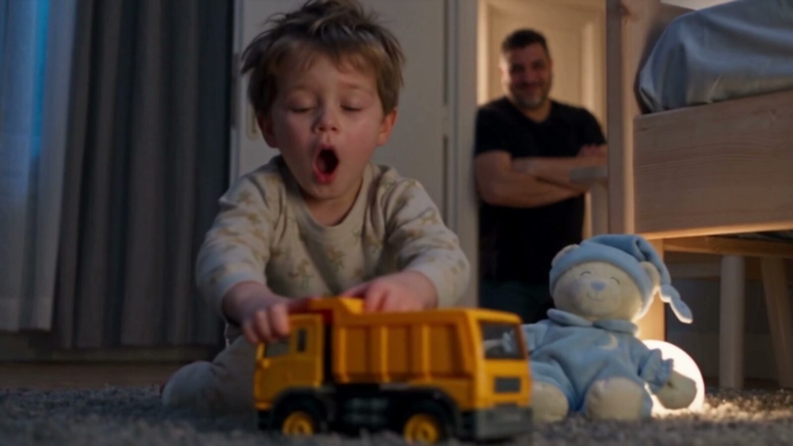 Le petit garçon bâille en jouant avec un camion jaune, son père le regarde depuis la porte, bras croisés