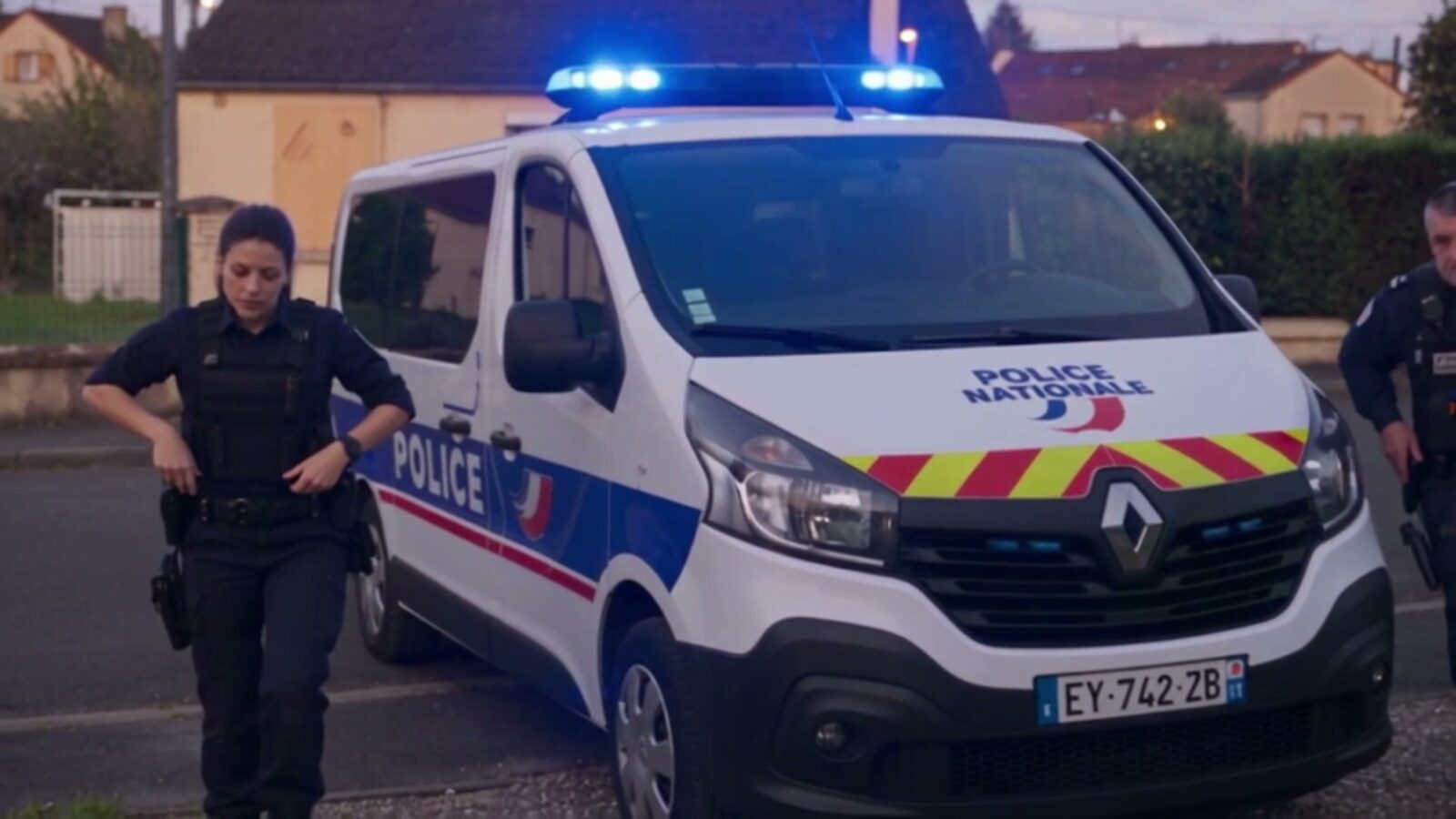 Un fourgon de police gyrophare allumé s'arrête devant la maison, une policière en descend