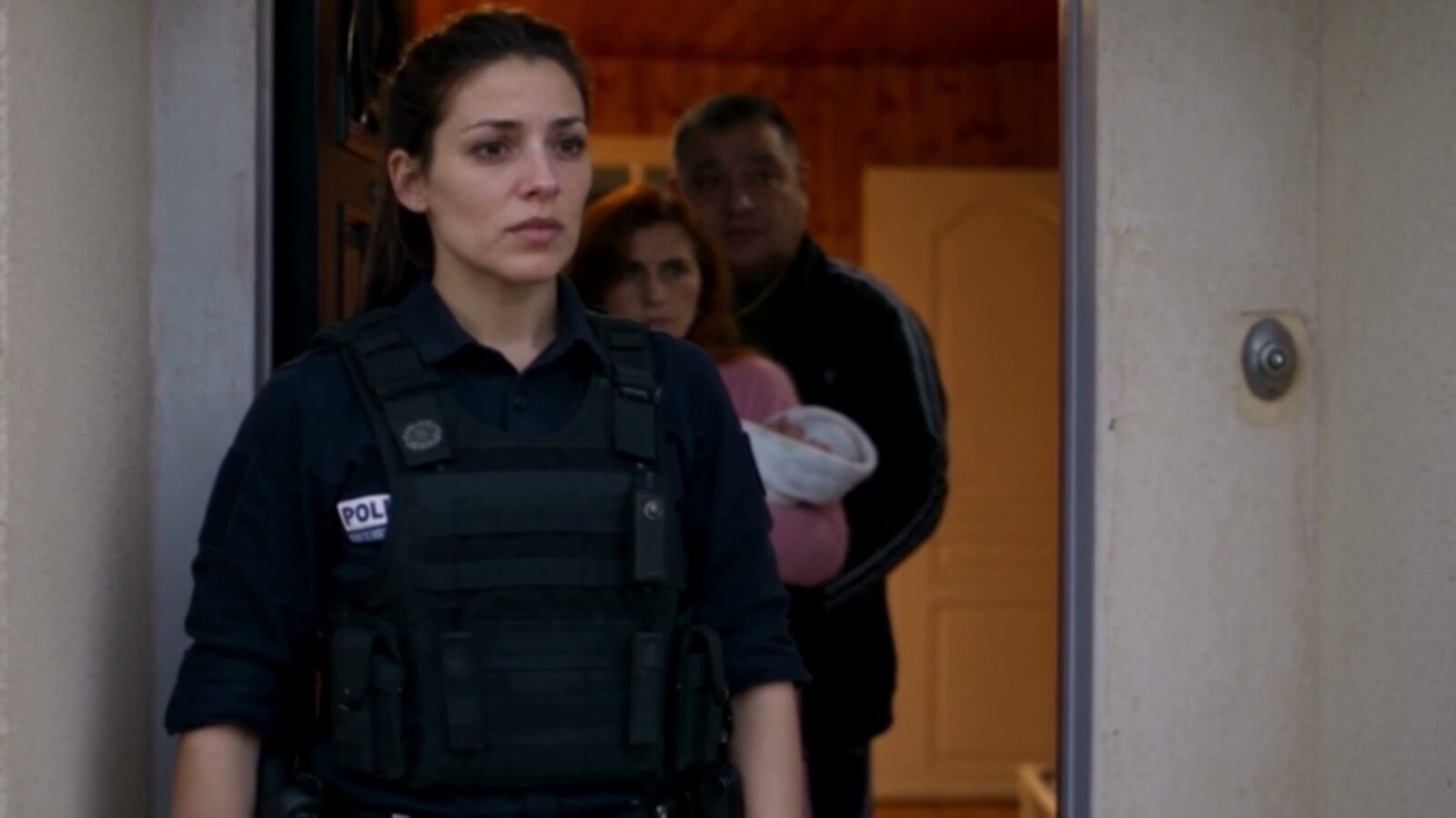 Une policière sur le pas de la porte, derrière elle les occupants de la maison, une femme tenant un bébé
