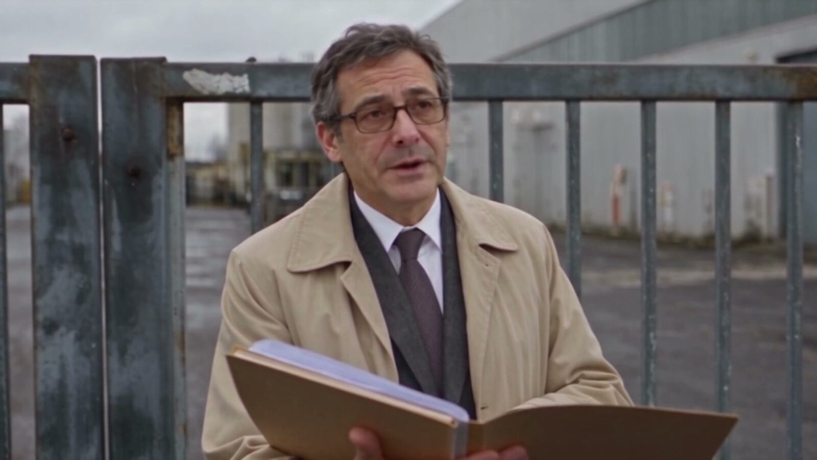 Un homme en imperméable, un dossier ouvert à la main, devant les grilles de l'usine