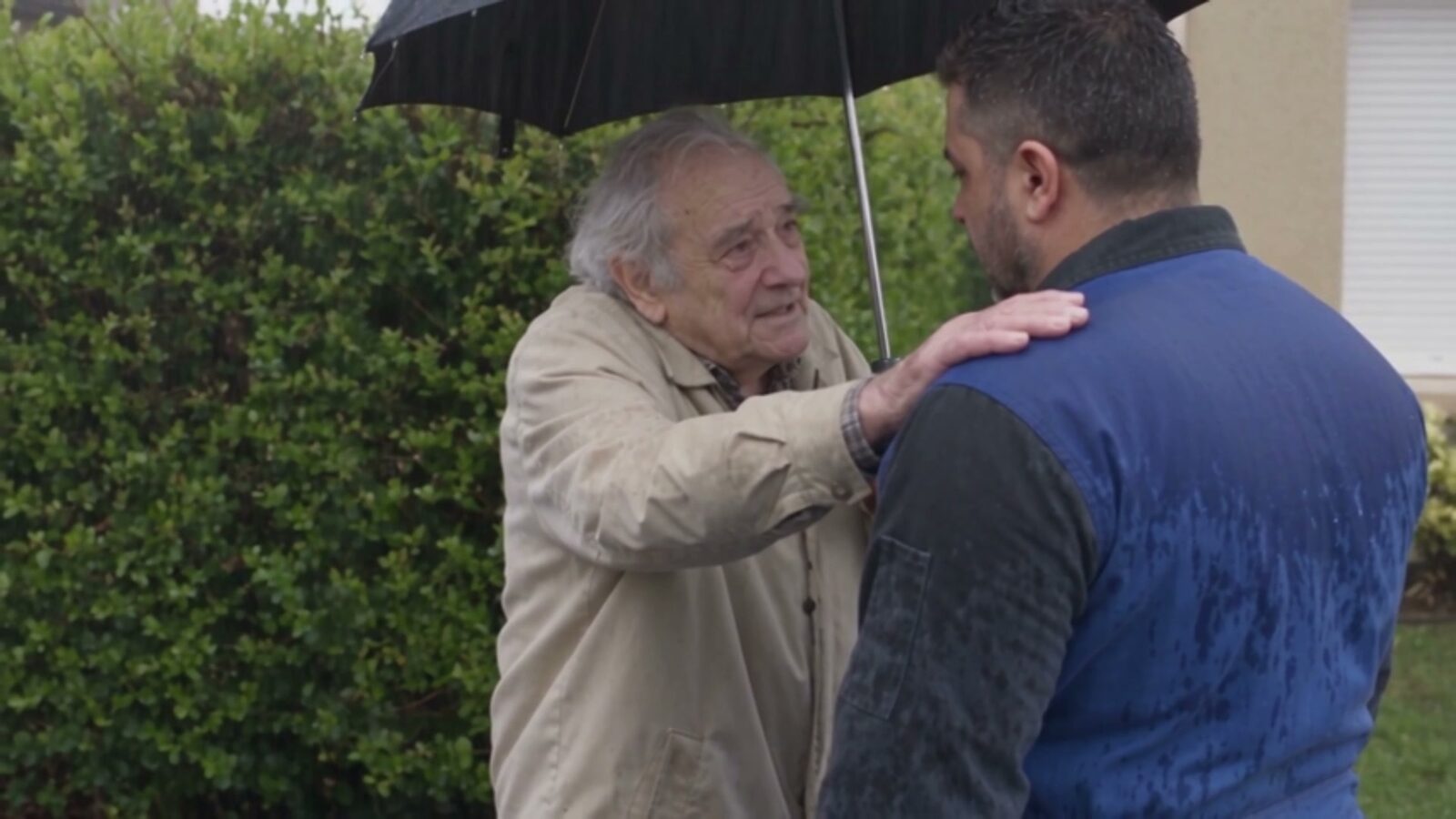 Sous la pluie, un vieil homme abrité par un parapluie pose la main sur l'épaule du père
