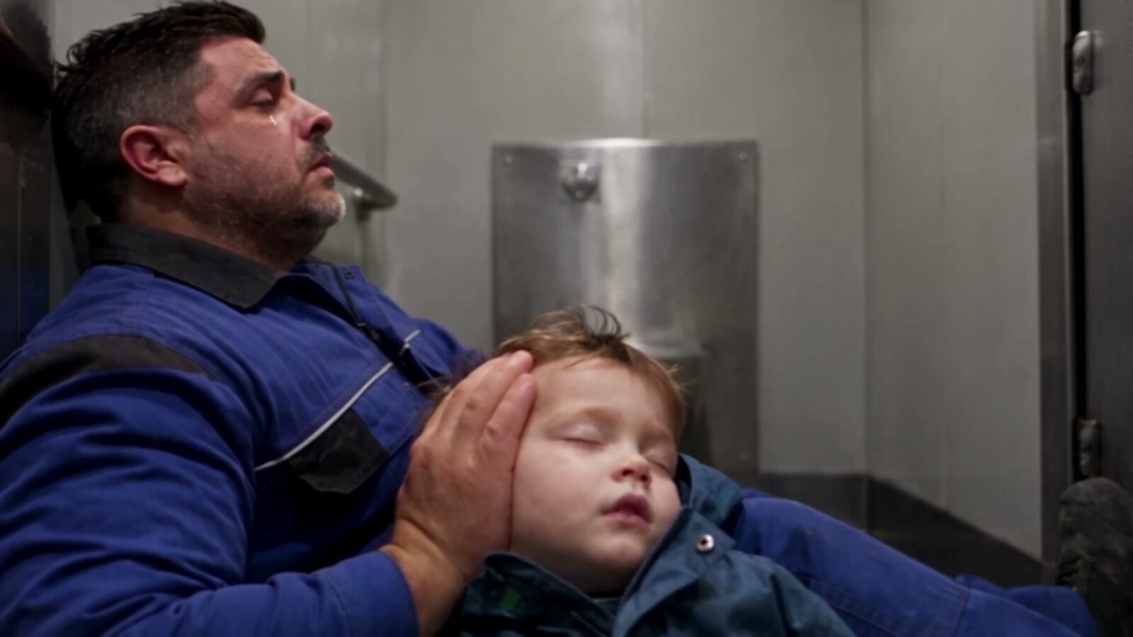 Le père en bleu de travail, adossé à une paroi en inox, pleure en tenant la tête de son fils endormi