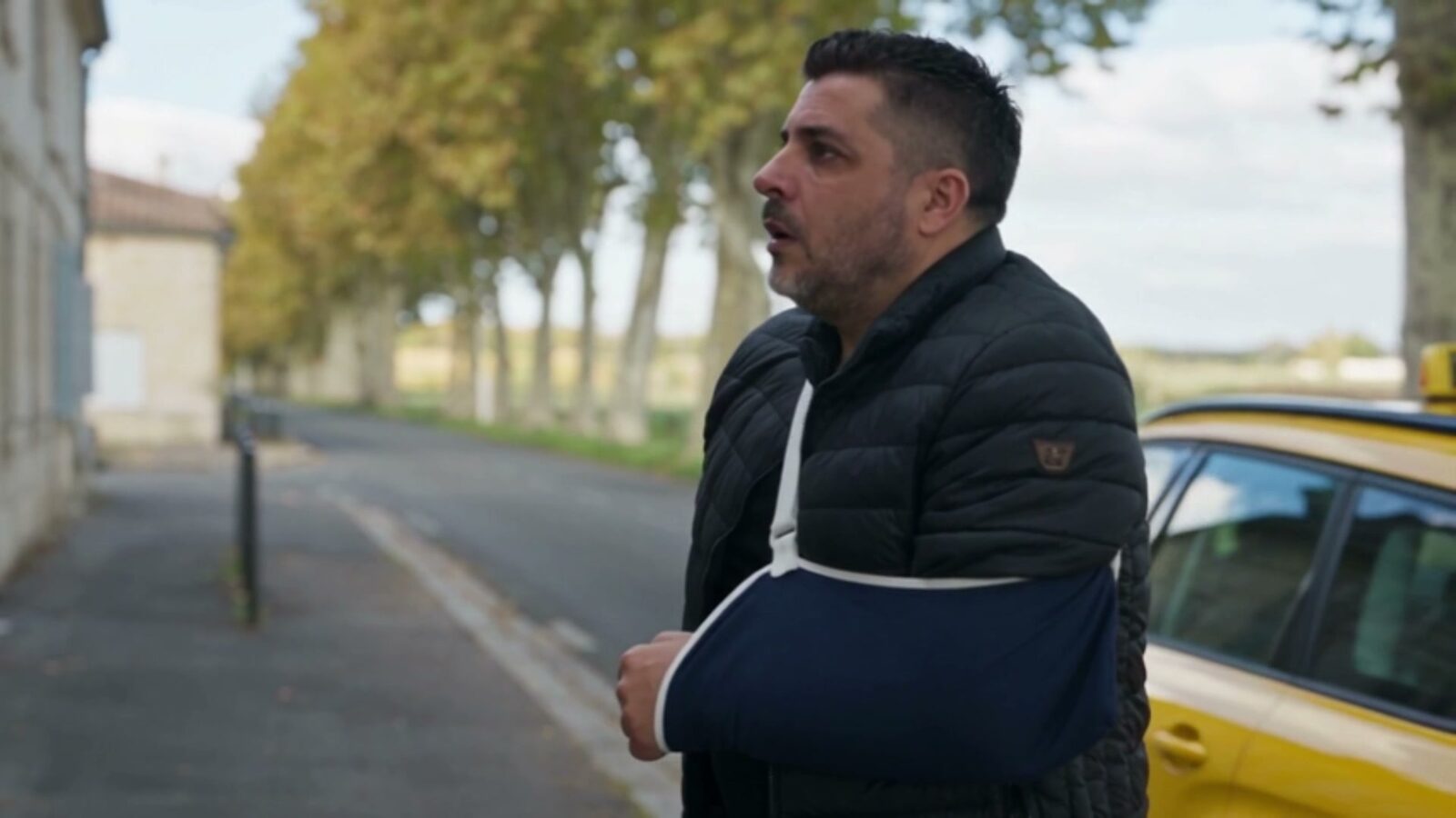 Le père, le bras en écharpe, dans une rue bordée d'arbres