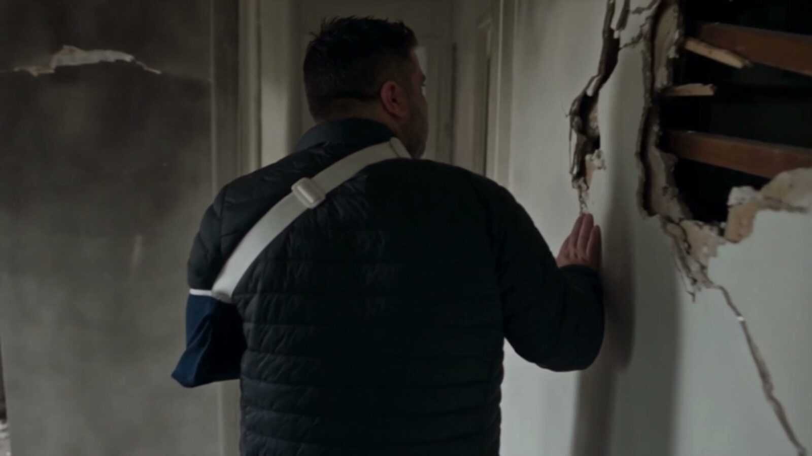 De dos, le bras en écharpe, le père pose la main sur un mur défoncé de sa maison