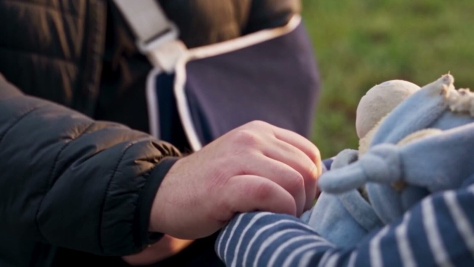 La main du père, bras en écharpe, près du doudou bleu que serre son fils