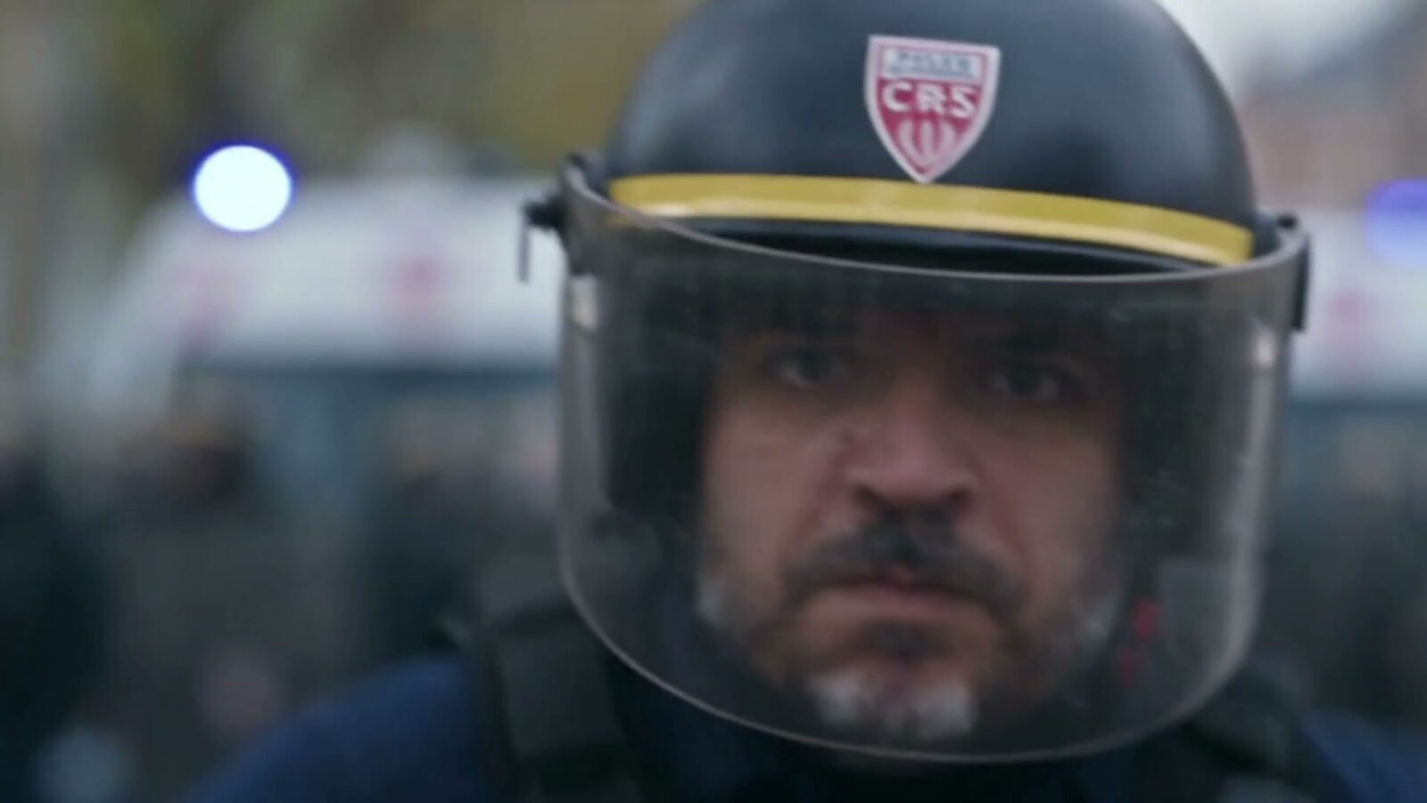 Le père sous son casque de CRS, visière baissée, gyrophare flou derrière lui